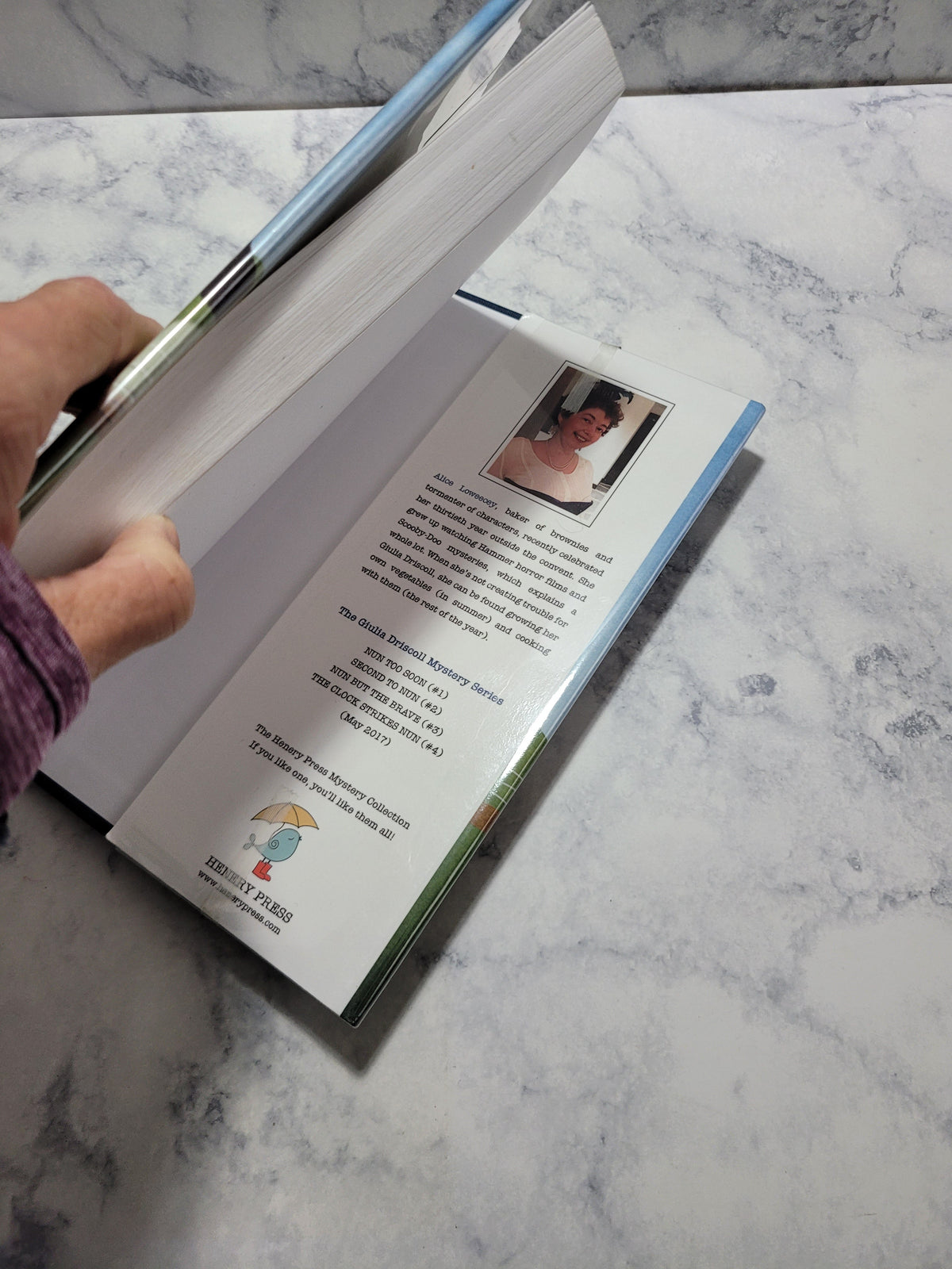 A person's hand and arm are visible as they open a printed booklet or book on a white and gray marble surface. The booklet pages are turned open, showing printed text and a small inset photo.