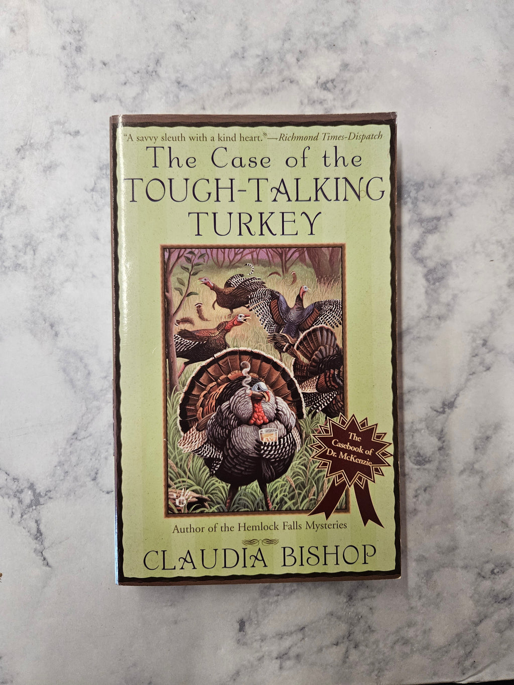 The book cover of 'The Case of the Tough-Talking Turkey' by Claudia Bishop, featuring a light green background and an illustration of a turkey, placed on a white and gray marble surface.
