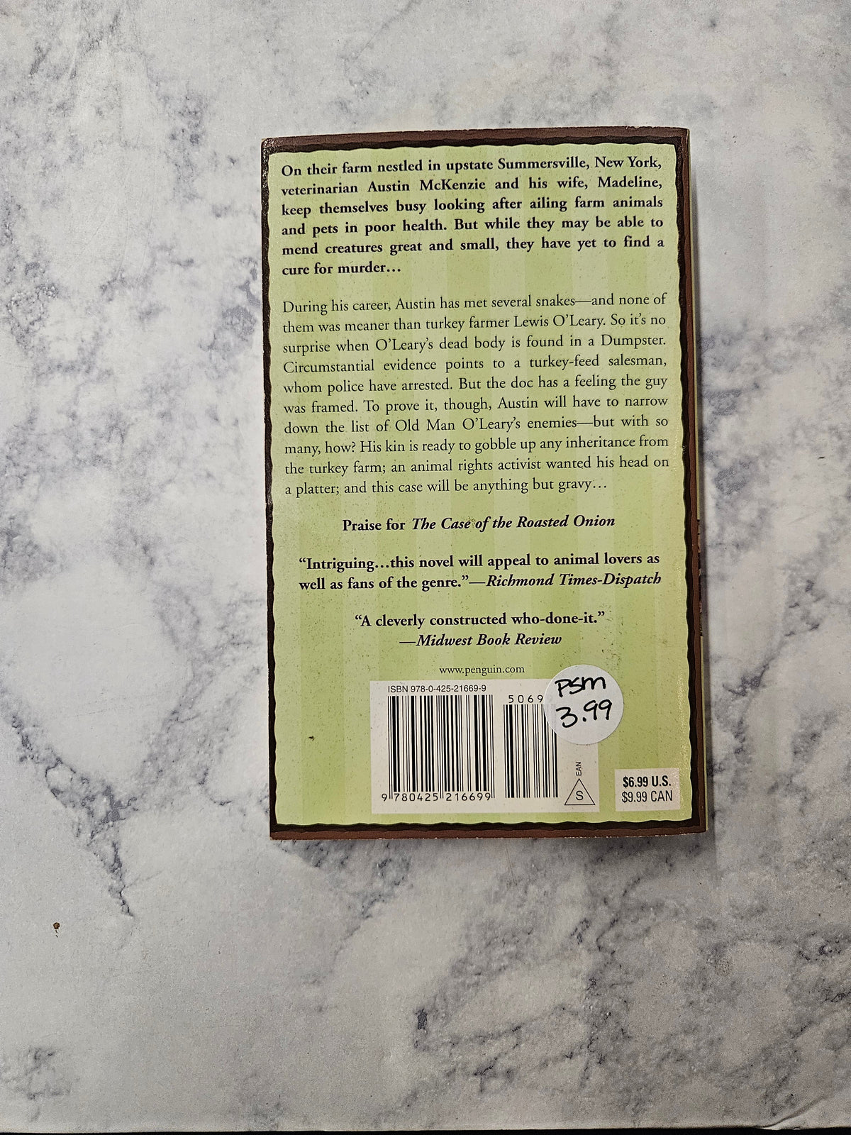 The back cover of a book lying on a white and gray marble surface. The cover has a light green background with black text, a barcode, and a handwritten price sticker that reads.