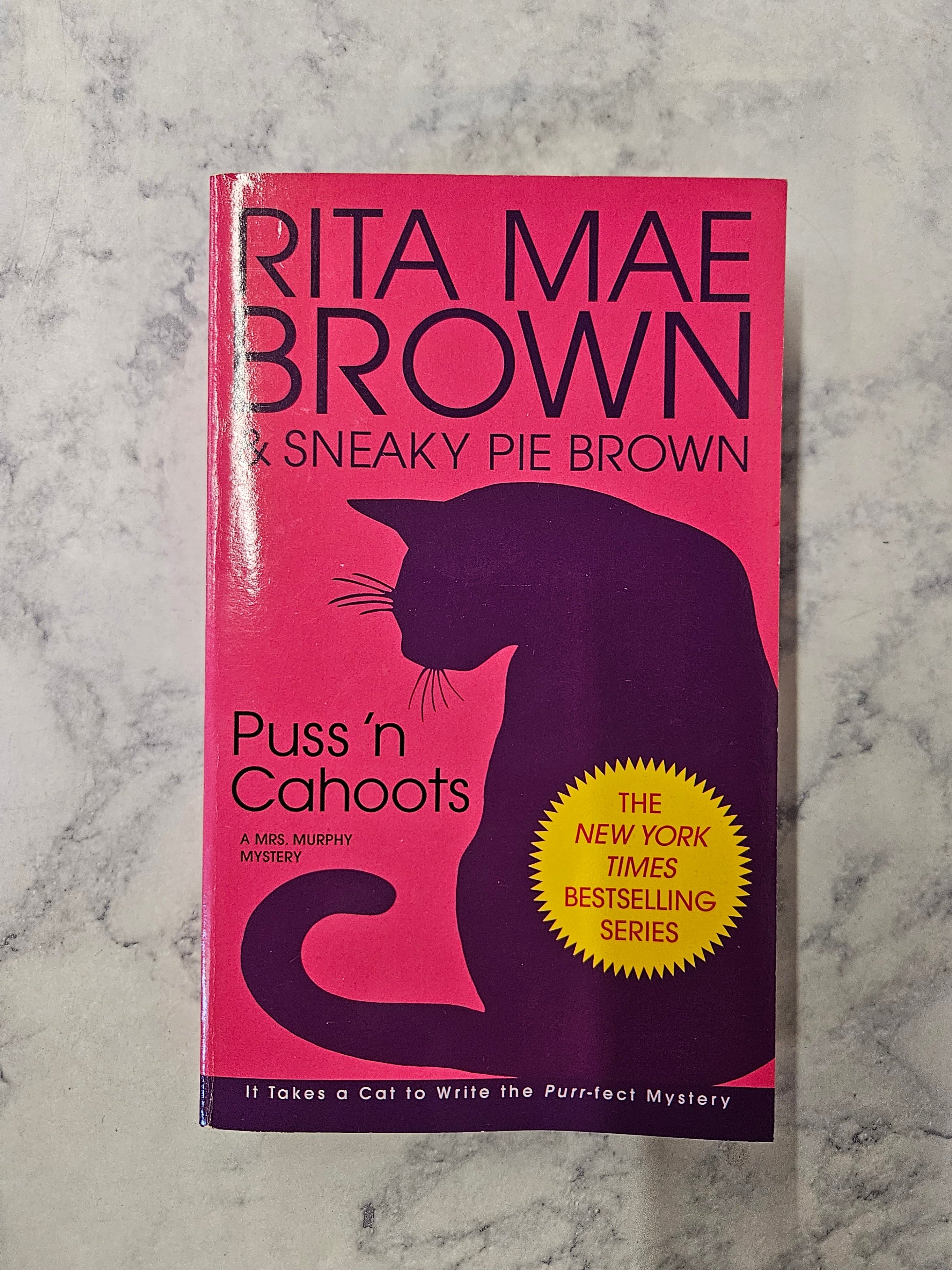 The book cover of 'Puss 'n Cahoots' by Rita Mae Brown & Sneaky Pie Brown, featuring a bright pink background with a dark purple cat silhouette and a yellow starburst badge, set against a white marble surface.