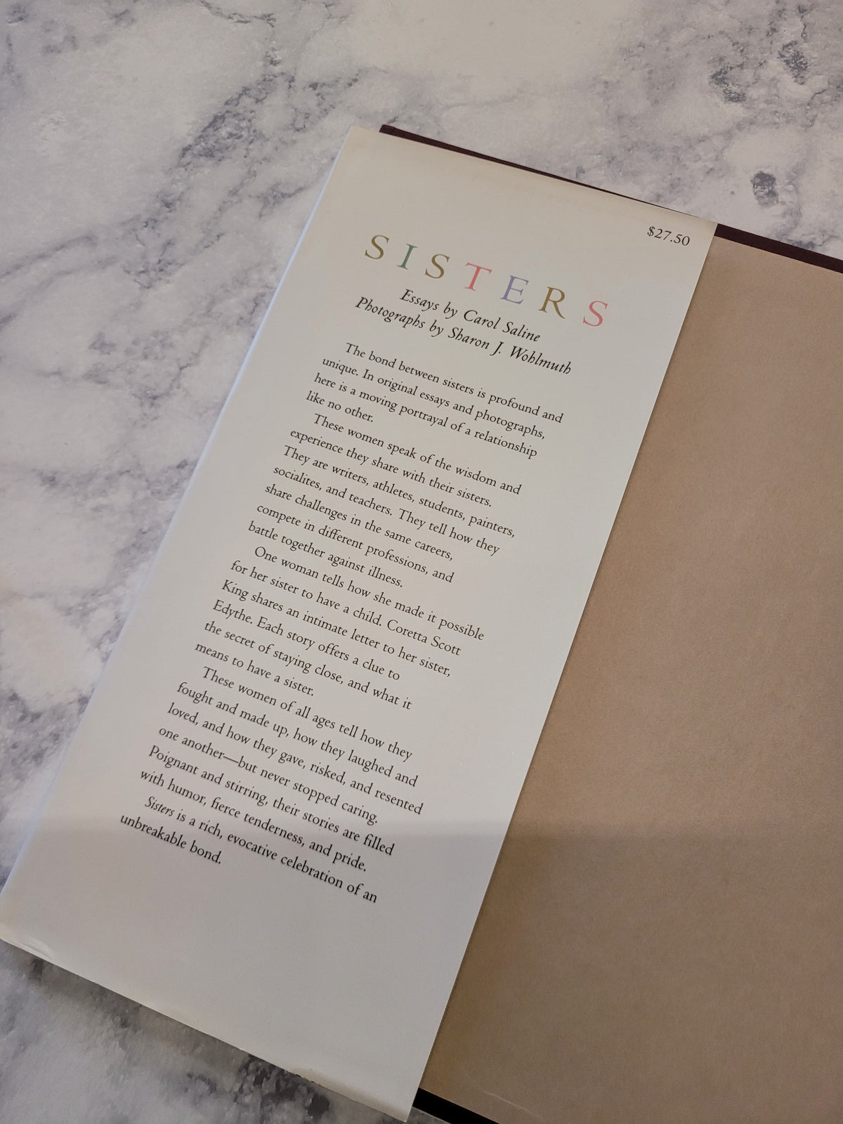 An open book or art print showing the interior page with the title 'SISTERS' and descriptive text, placed on a light marble surface next to a brown box or folder.