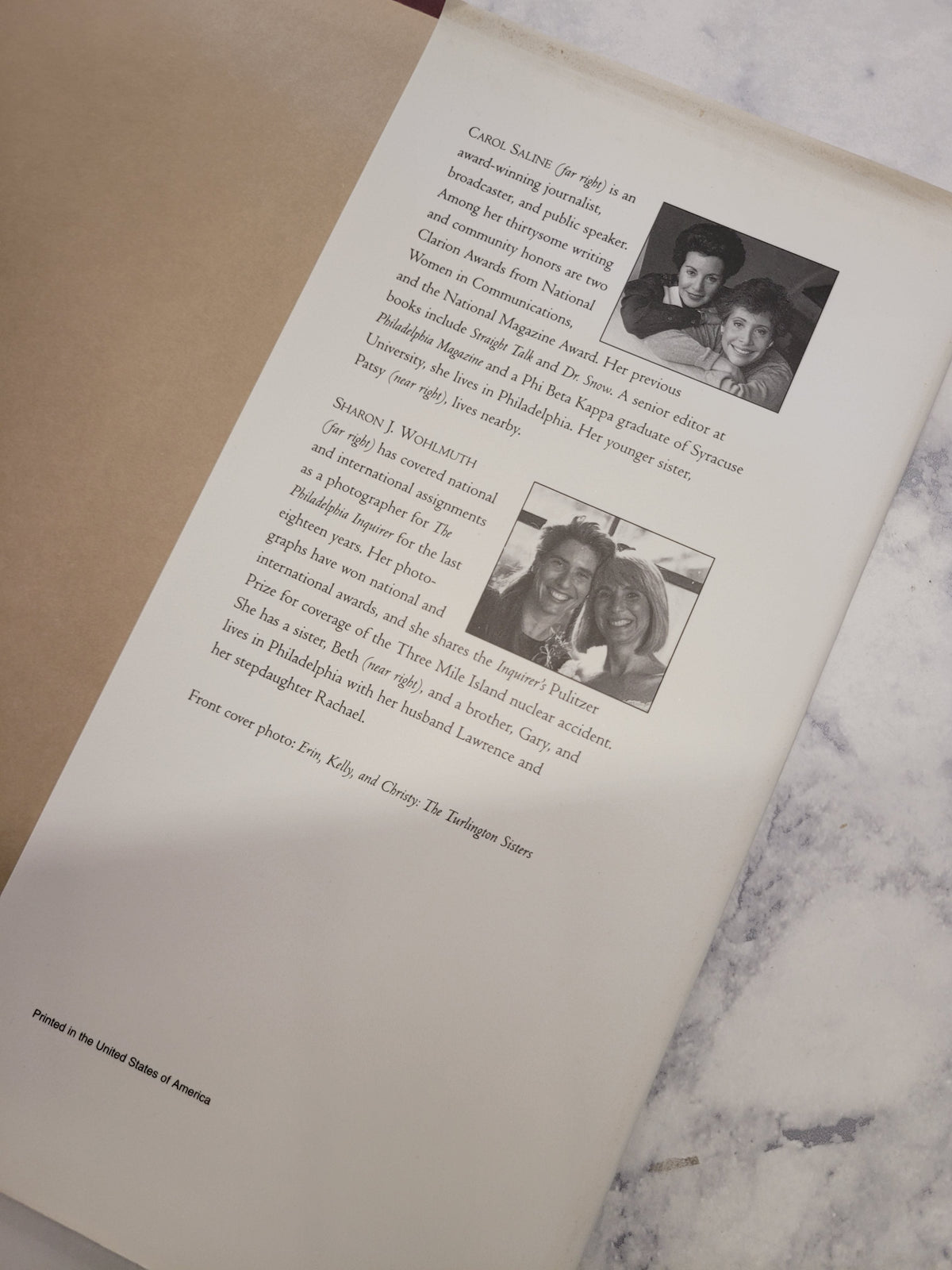 An open book or booklet showing printed pages with two small black-and-white photographs and blocks of text, photographed on a marble-like surface with a tan page or folder visible underneath.