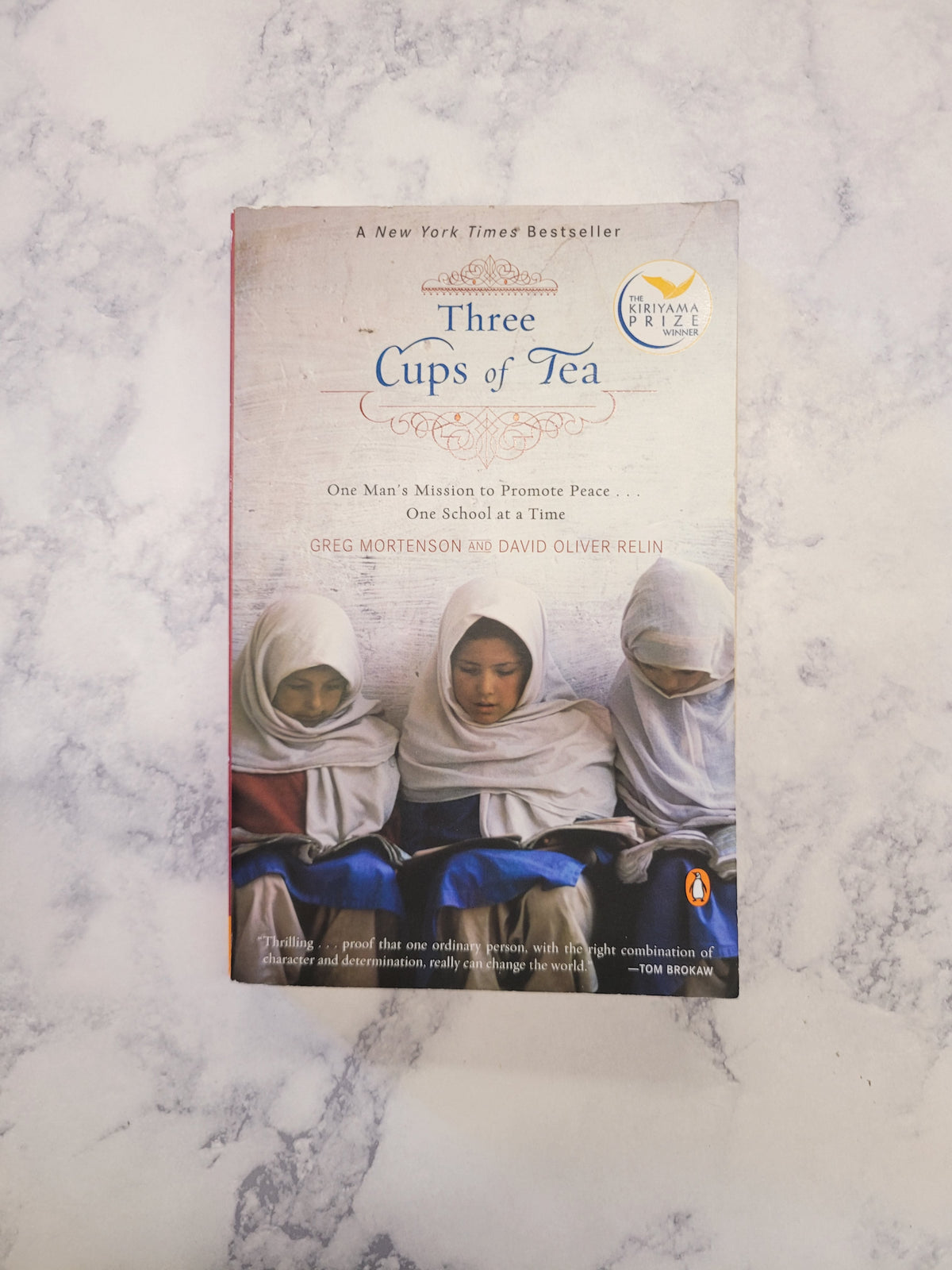 The book 'Three Cups of Tea' by Poul Nørgaard lies flat on a white and gray marble-patterned surface. The cover features a photograph of three women in white hijabs and displays the Penguin logo in the top right corner.