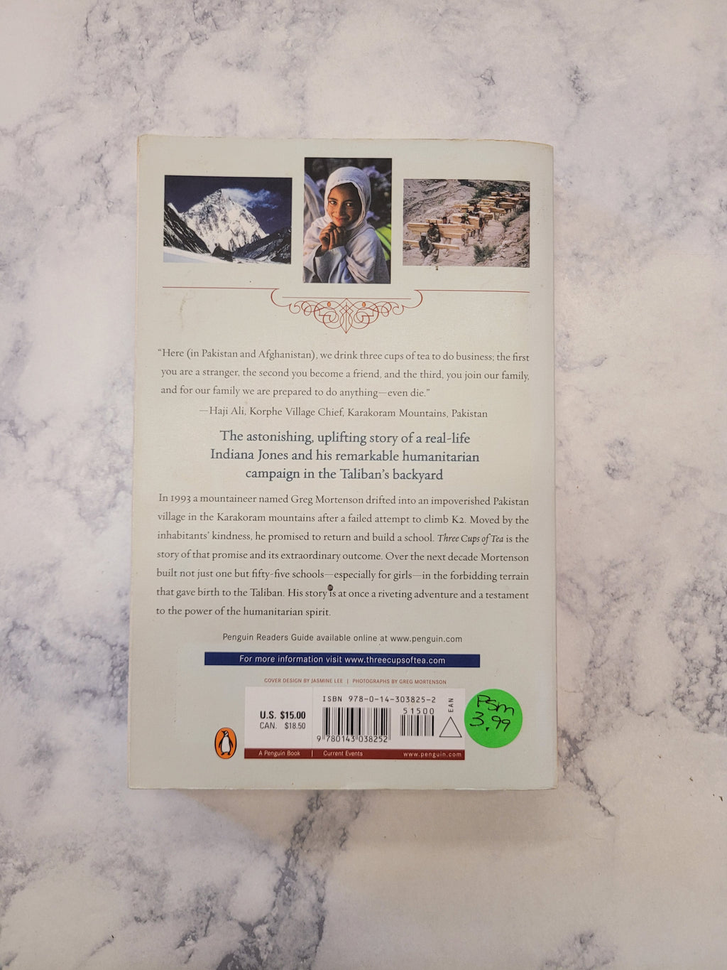 The back cover of a book displayed flat on a white and gray marble surface. The cover features three small illustrations, a barcode, and a green circular sticker.