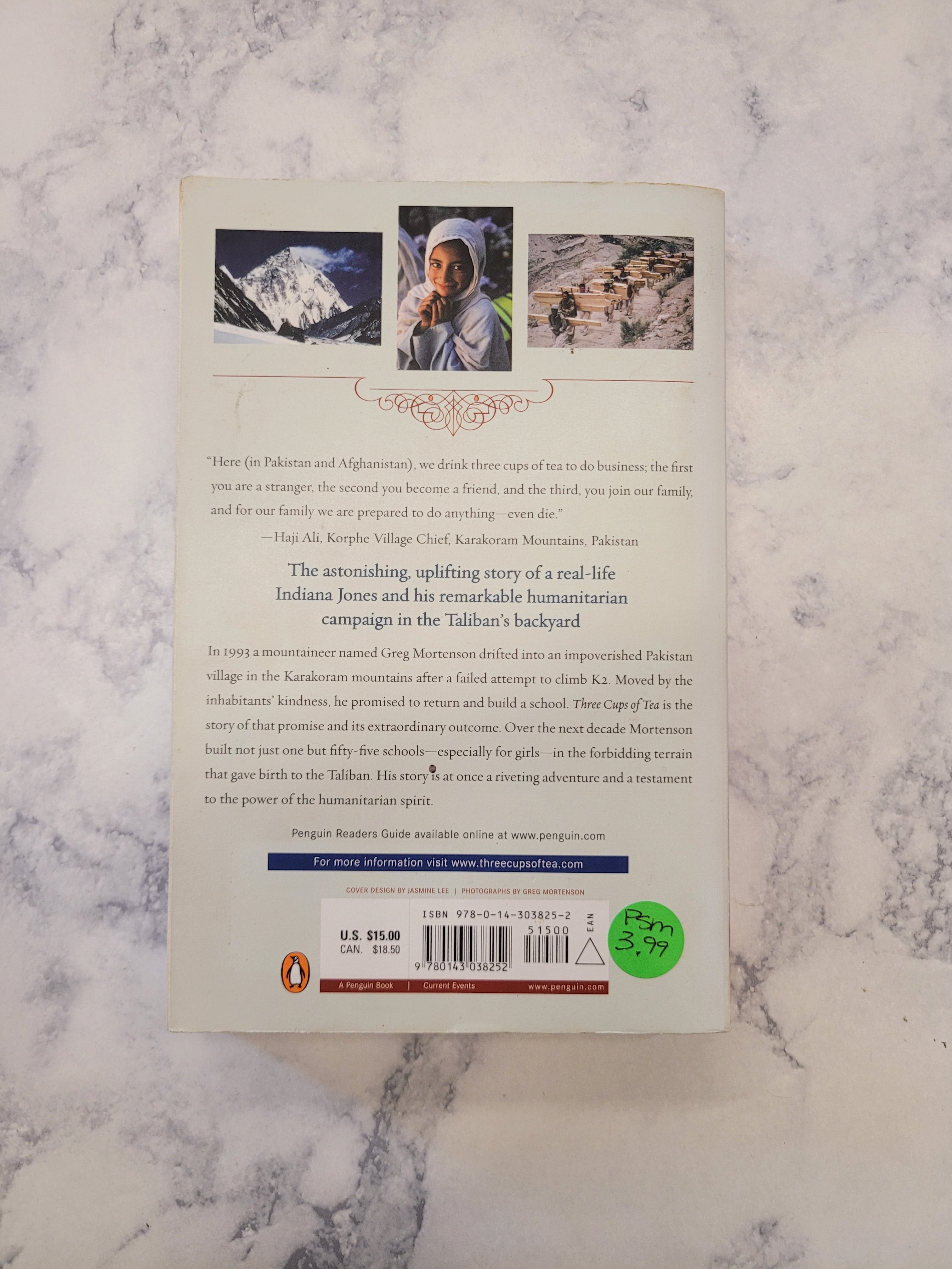 The back cover of a book displayed flat on a white and gray marble surface. The cover features three small illustrations, a barcode, and a green circular sticker.