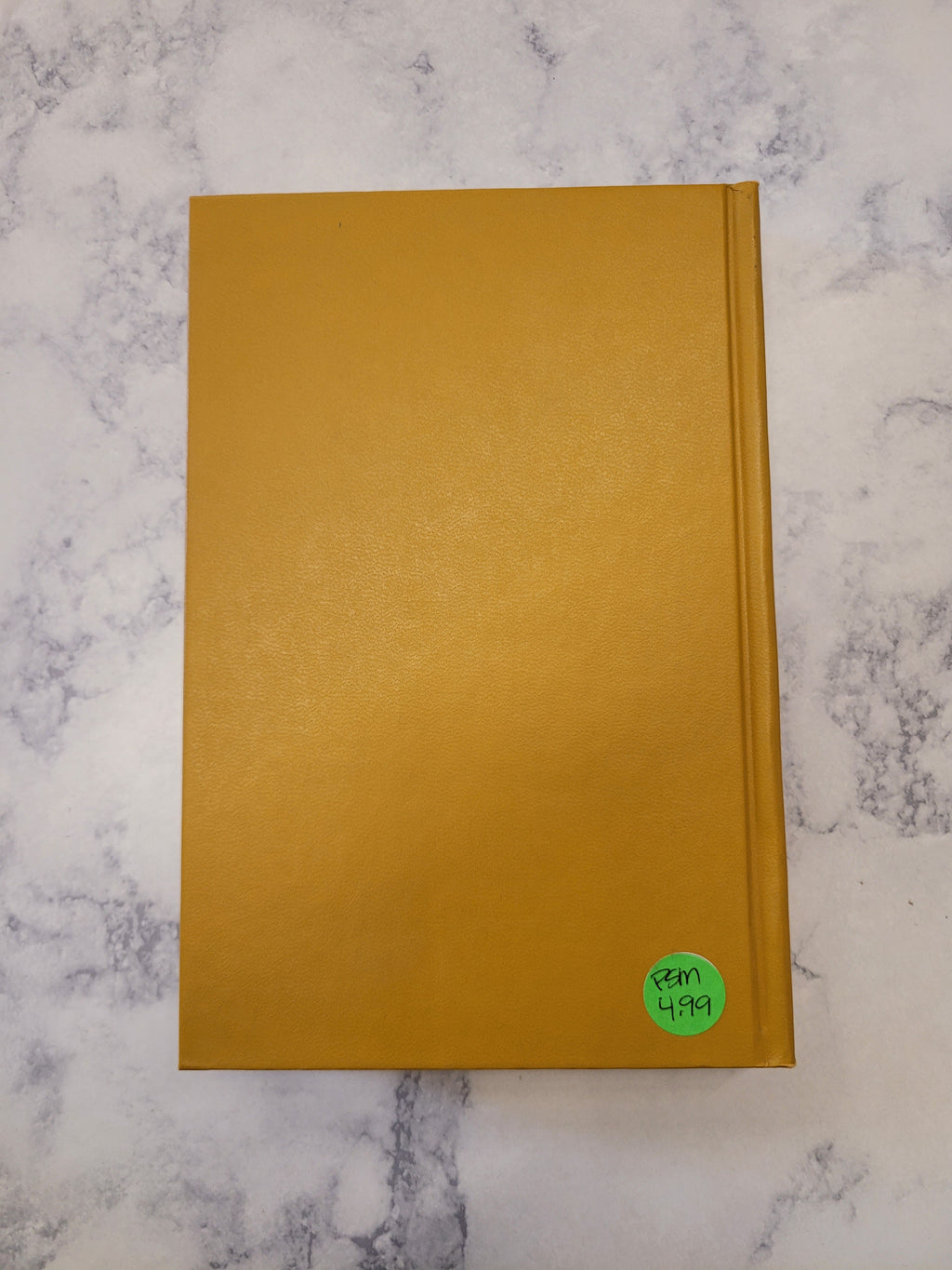 A mustard yellow textured hardcover notebook or book lying flat on a white and gray marble-patterned surface, with a small green circular sticker on the top right corner.