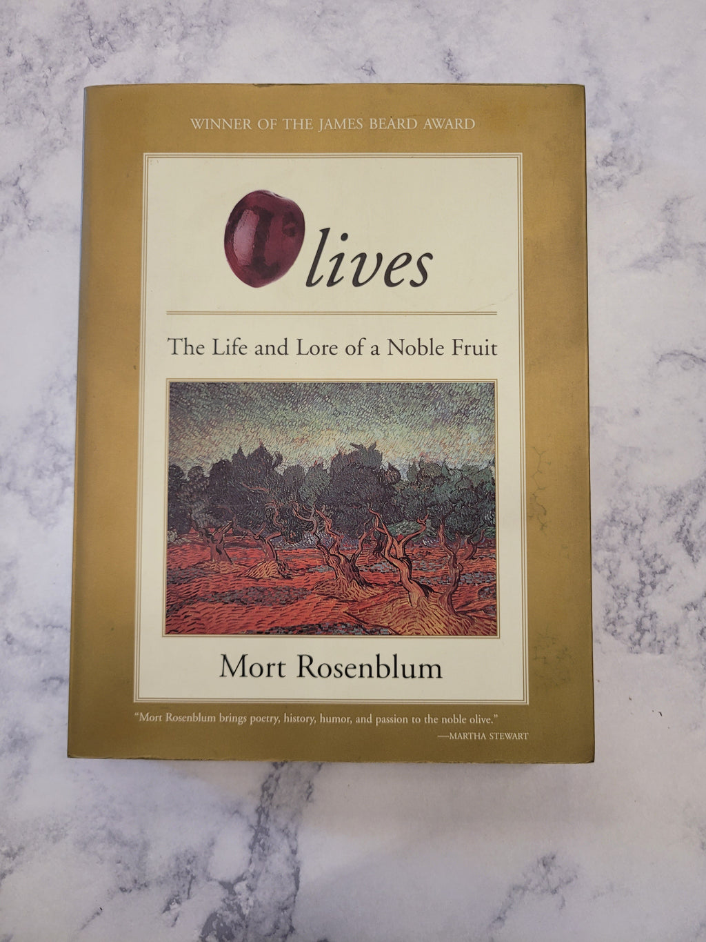 The book 'Olives: The Life and Lore of a Noble Fruit' by Mort Rosenblum, featuring a gold-colored cover with a cream central panel, a red apple illustration, and a landscape painting, displayed on a white marble surface.