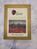 The book 'Olives: The Life and Lore of a Noble Fruit' by Mort Rosenblum, featuring a gold-colored cover with a cream central panel, a red apple illustration, and a landscape painting, displayed on a white marble surface.