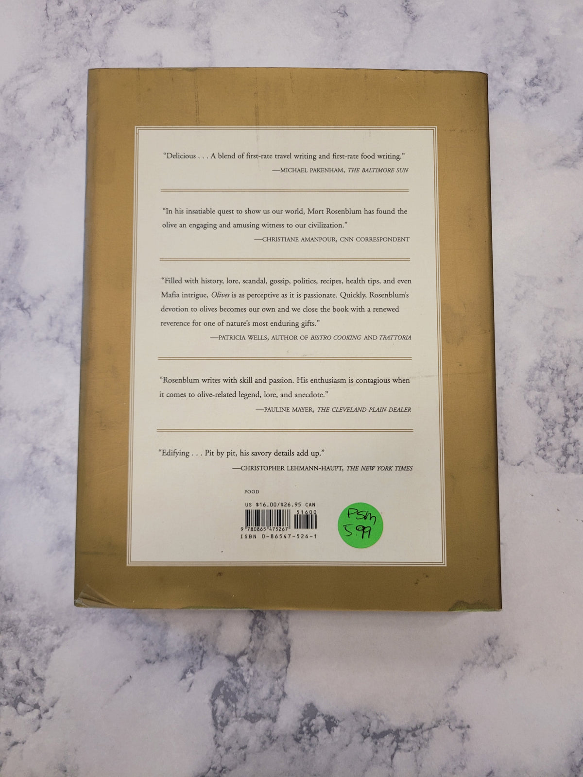 The back cover of a book with a tan background and a large cream-colored text panel, placed on a white and gray marble surface. A green circular sticker is visible on the right side of the panel.