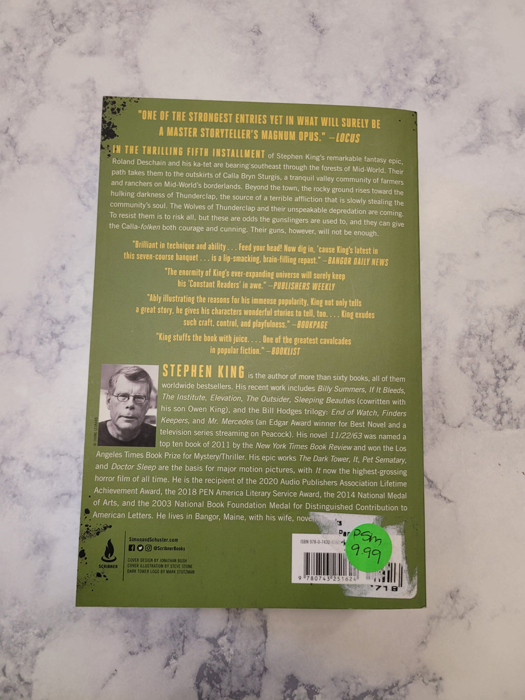 The back cover of the book 'The Wolves of Calla' by Stephen King, featuring a sage green background with yellow text, a barcode, and a small black-and-white portrait, photographed on a white and gray marble surface.