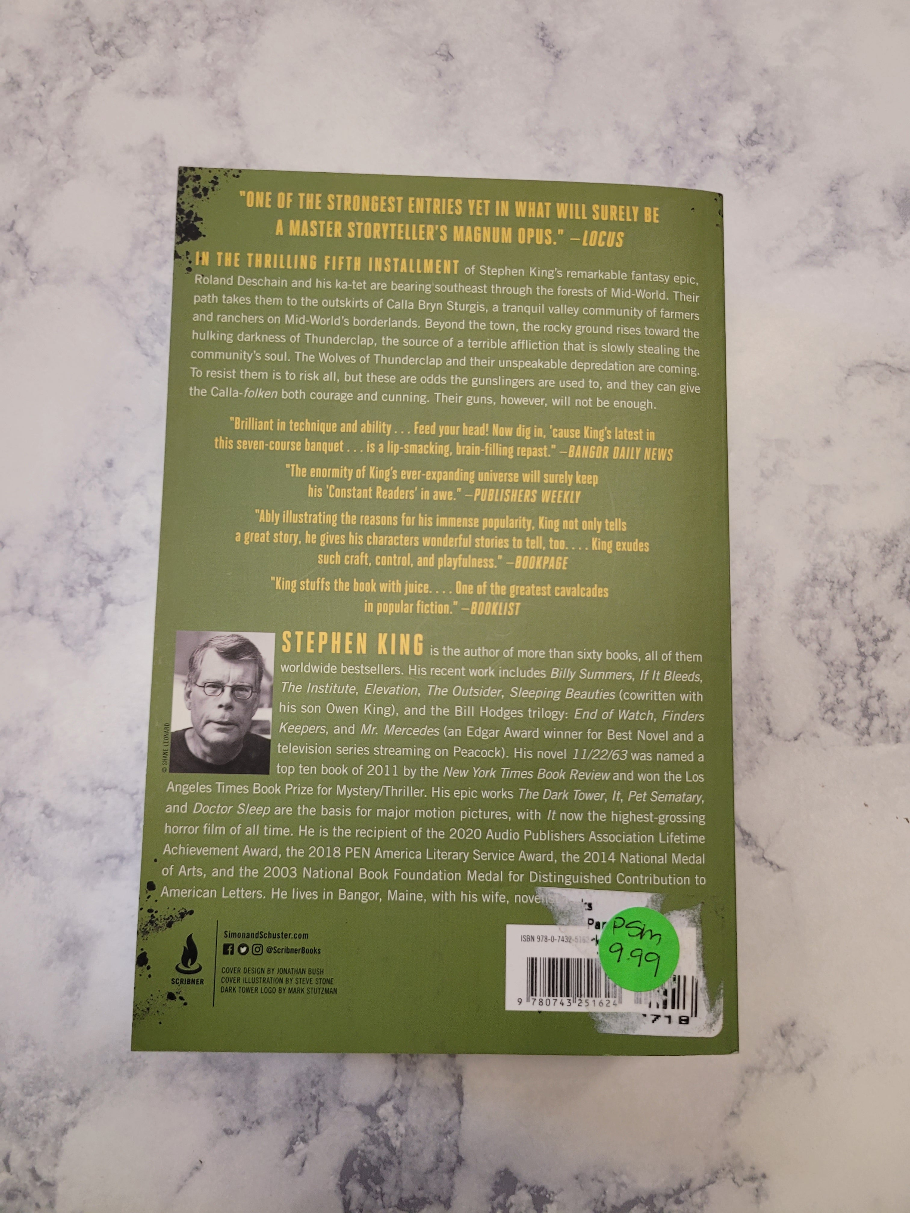 The back cover of the book 'The Wolves of Calla' by Stephen King, featuring a sage green background with yellow text, a barcode, and a small black-and-white portrait, photographed on a white and gray marble surface.