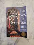 The book cover of 'The Widows of Malabar Hill' by Sujata Massey, featuring a purple background with white title text and an ornate Moorish arch illustration, placed on a light gray marble surface.