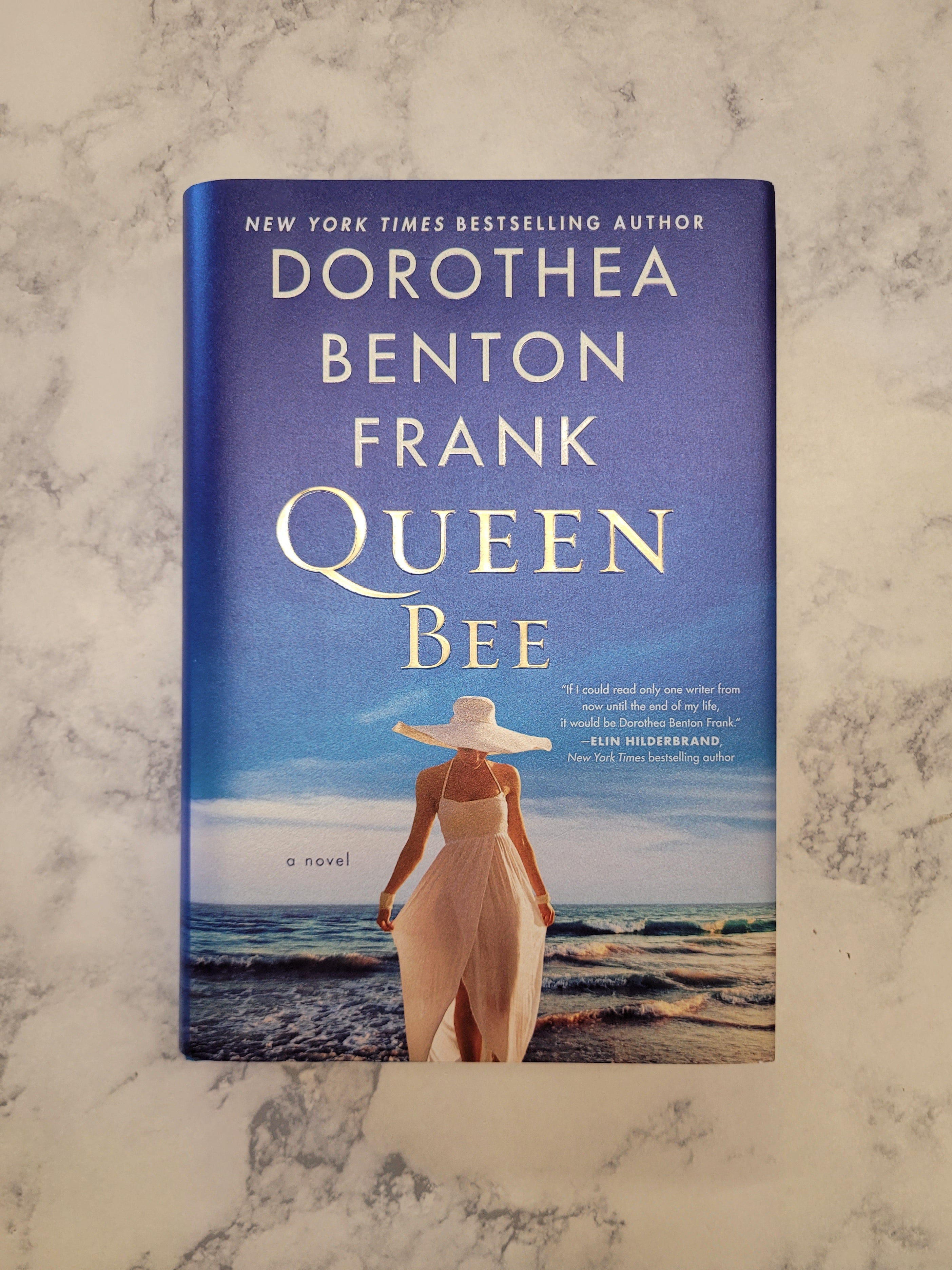 The book cover of 'Queen Bee' by Dorothea Benton Frank is displayed flat on a light gray and white marble surface. The cover features a blue sky and ocean background with a woman in a white dress and hat standing on the beach.