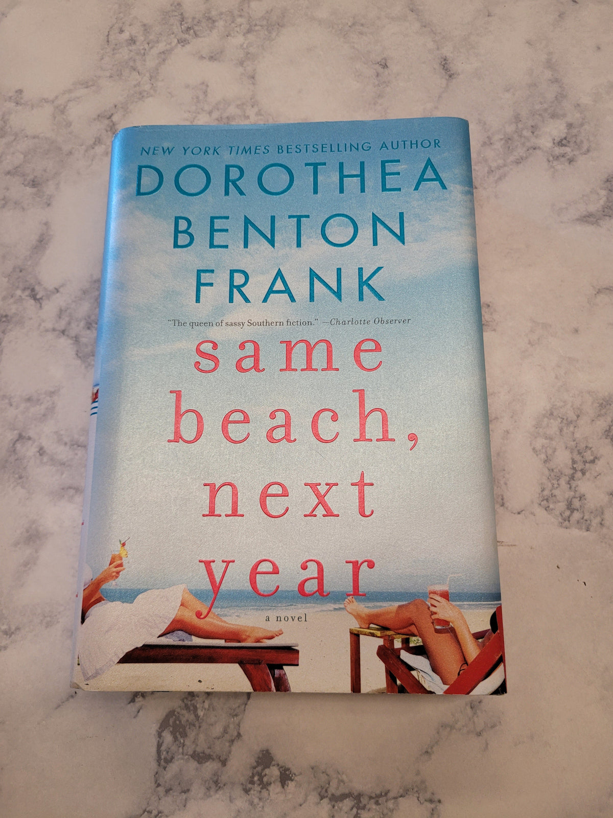 The book cover of 'Same Beach, Next Year' by Dorothea Benton Frank, placed on a light gray and white marble surface. The cover features a light blue sky background with red text and illustrations of people on a beach.