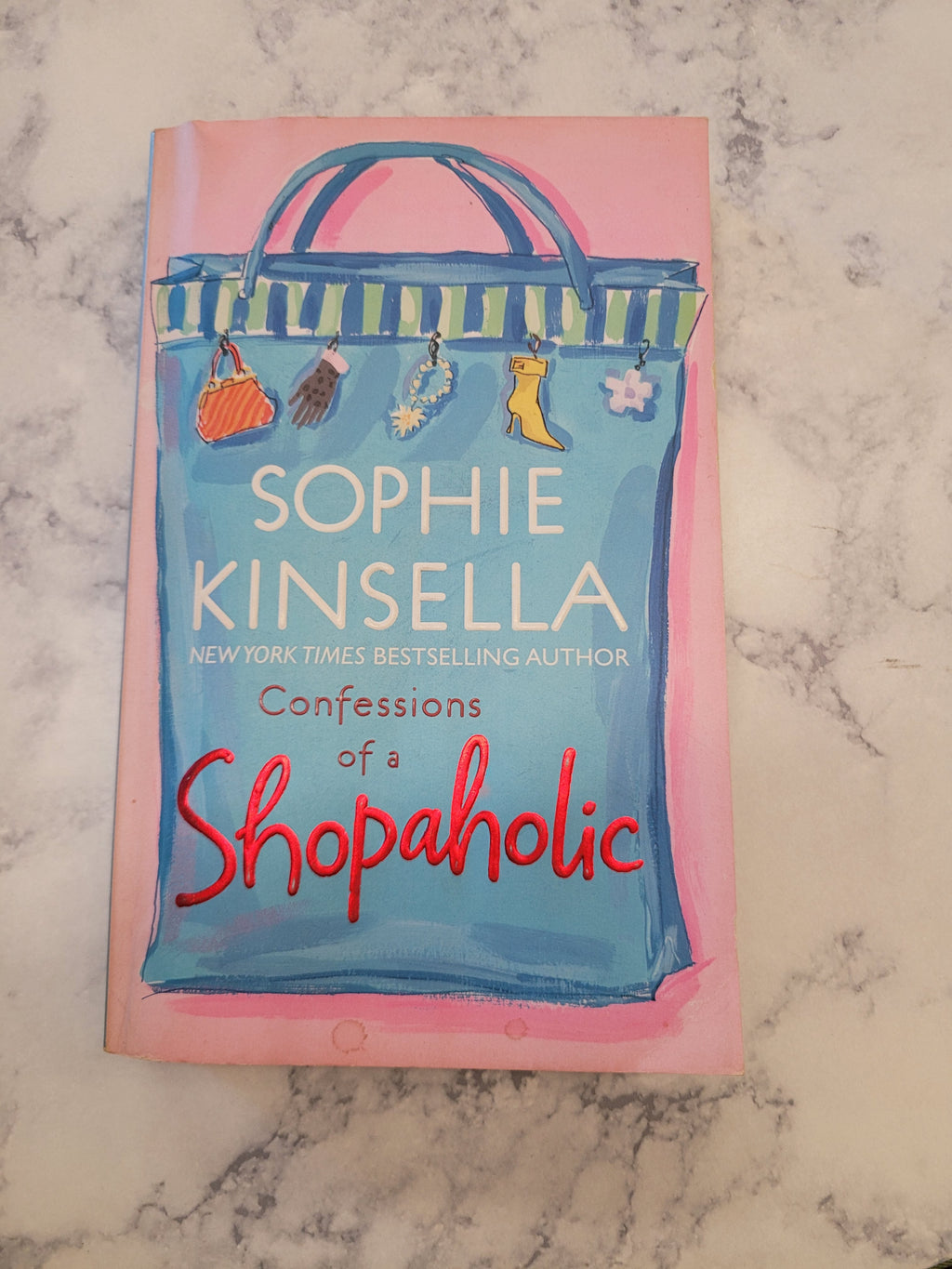 The book cover of 'Confessions of a Shopaholic' by Sophie Kinsella, featuring a pink border and a blue shopping bag illustration, placed on a white and gray marble surface.