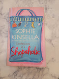 The book cover of 'Confessions of a Shopaholic' by Sophie Kinsella, featuring a pink border and a blue shopping bag illustration, placed on a white and gray marble surface.