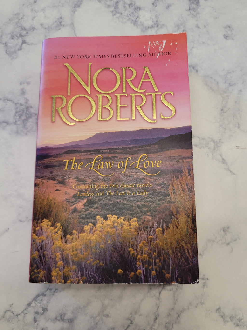 The book 'The Law of Love' by Nora Roberts, featuring a pink-to-purple sunset landscape cover with gold lettering, placed on a white and gray marble surface.
