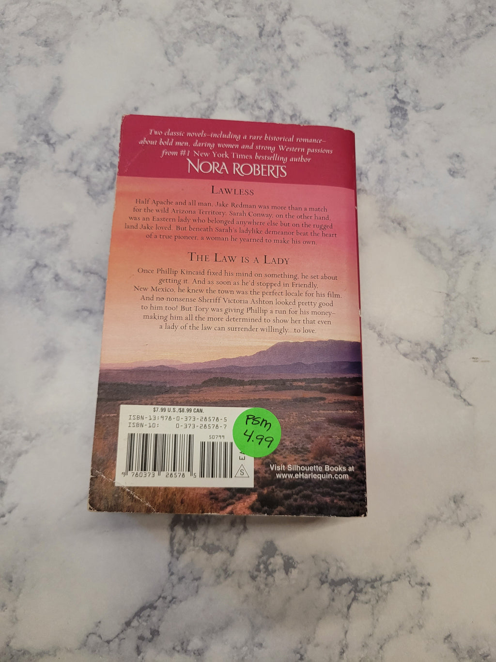The back cover of a book by Nora Roberts, featuring a scenic sunset landscape and a green price sticker, displayed on a white and gray marble surface.