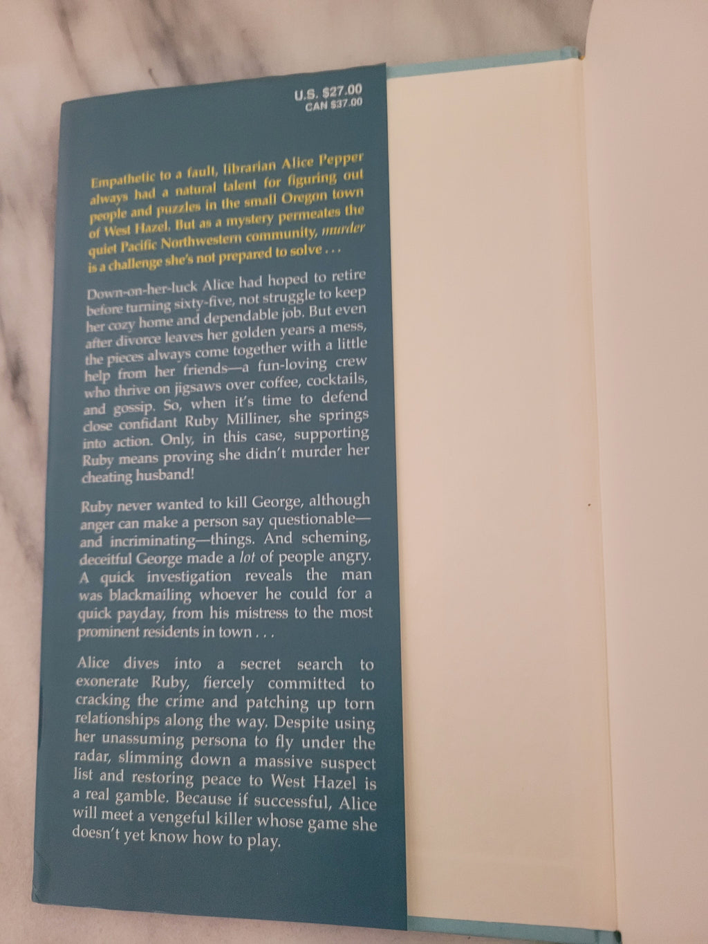 A book or booklet shown open, with a dark teal cover featuring white and yellow text, and blank cream pages visible underneath on a light marble-like surface.