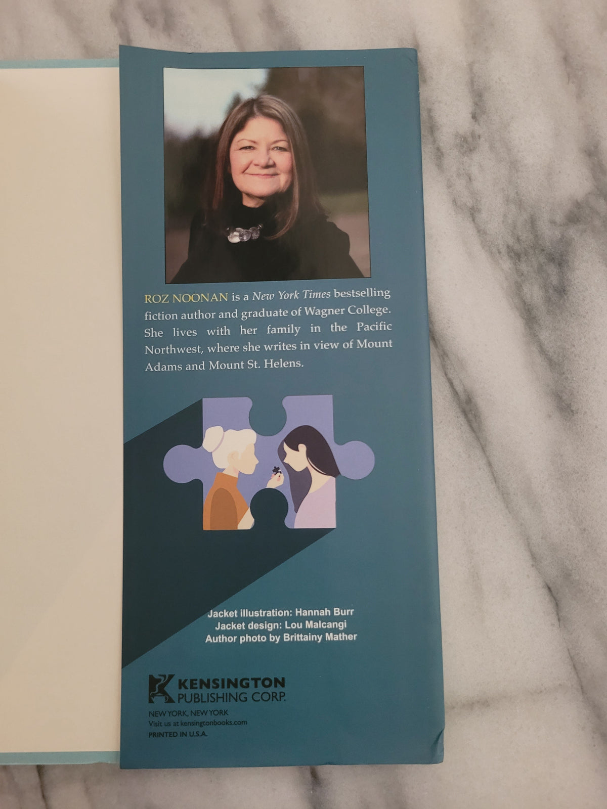 A folded teal and navy printed brochure or invitation card with text, a small puzzle-piece illustration, and a portrait photo of a woman, placed on a white marble surface.