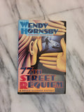 The book cover of '77th Street Requiem' by Wendy Hornsby, featuring a stylized illustration of hands and a dark figure, photographed on a white and gray marble surface.