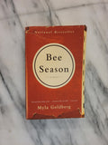 A vintage copy of the book 'Bee Season' by Myla Goldberg, featuring a rust-orange cover with a cream-colored circular logo, placed on a white marble surface.
