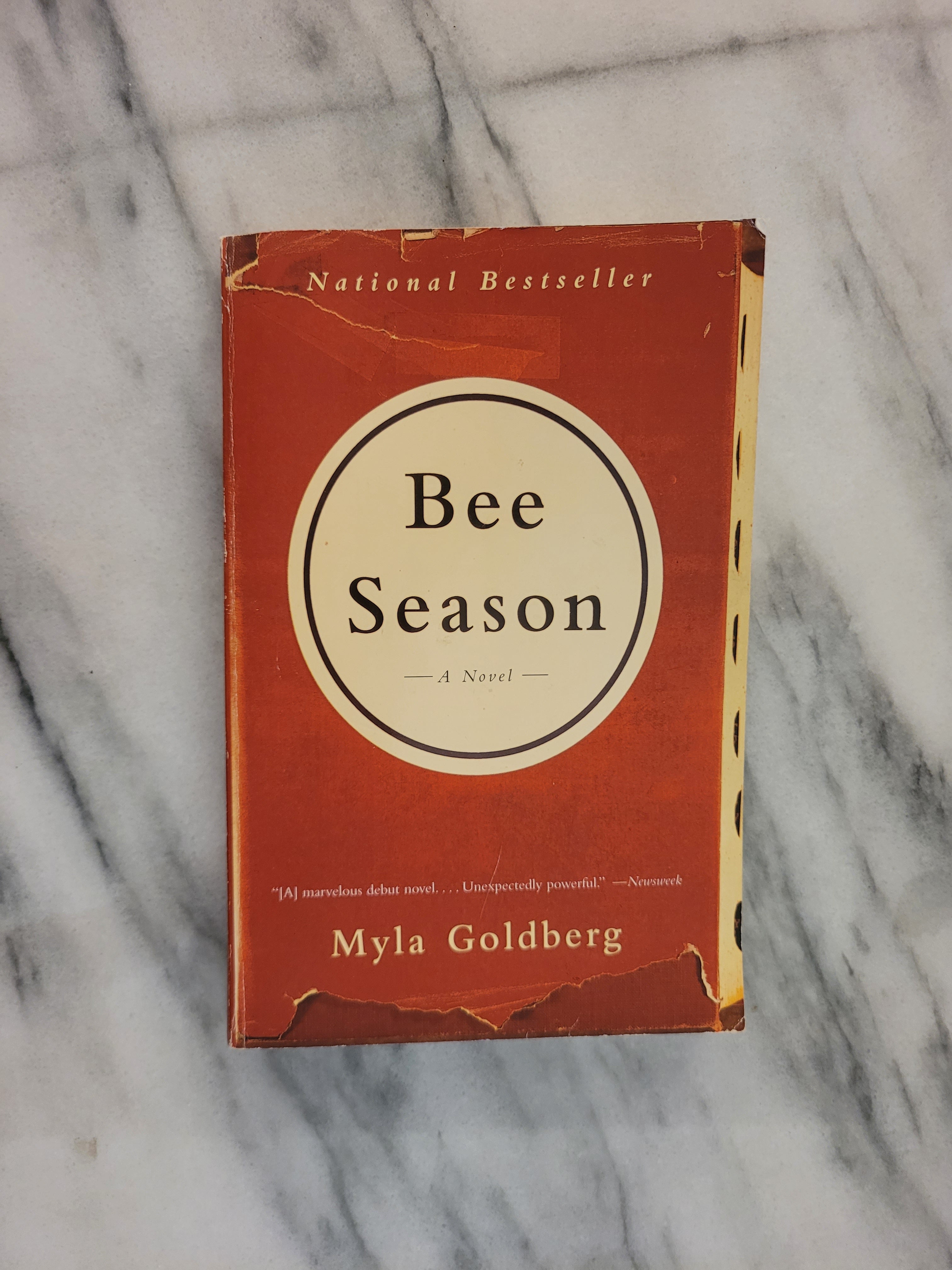 A vintage copy of the book 'Bee Season' by Myla Goldberg, featuring a rust-orange cover with a cream-colored circular logo, placed on a white marble surface.