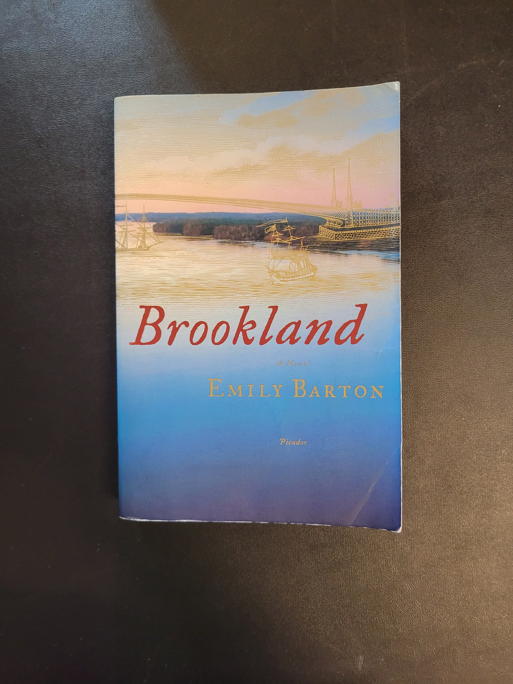 The book 'Brookland' by Emily Barton, published by Faber & Faber, shown flat on a dark surface. The cover features a scenic photograph of a pier at sunset with blue, yellow, and pink tones, and the title is written in red script.