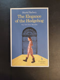 The book cover of 'The Elegance of the Hedgehog' by Muriel Barbery, featuring a blue background, a yellow diagonal section, and an illustration of a girl in a pink plaid dress standing in front of a large door, placed on a dark textured surface.