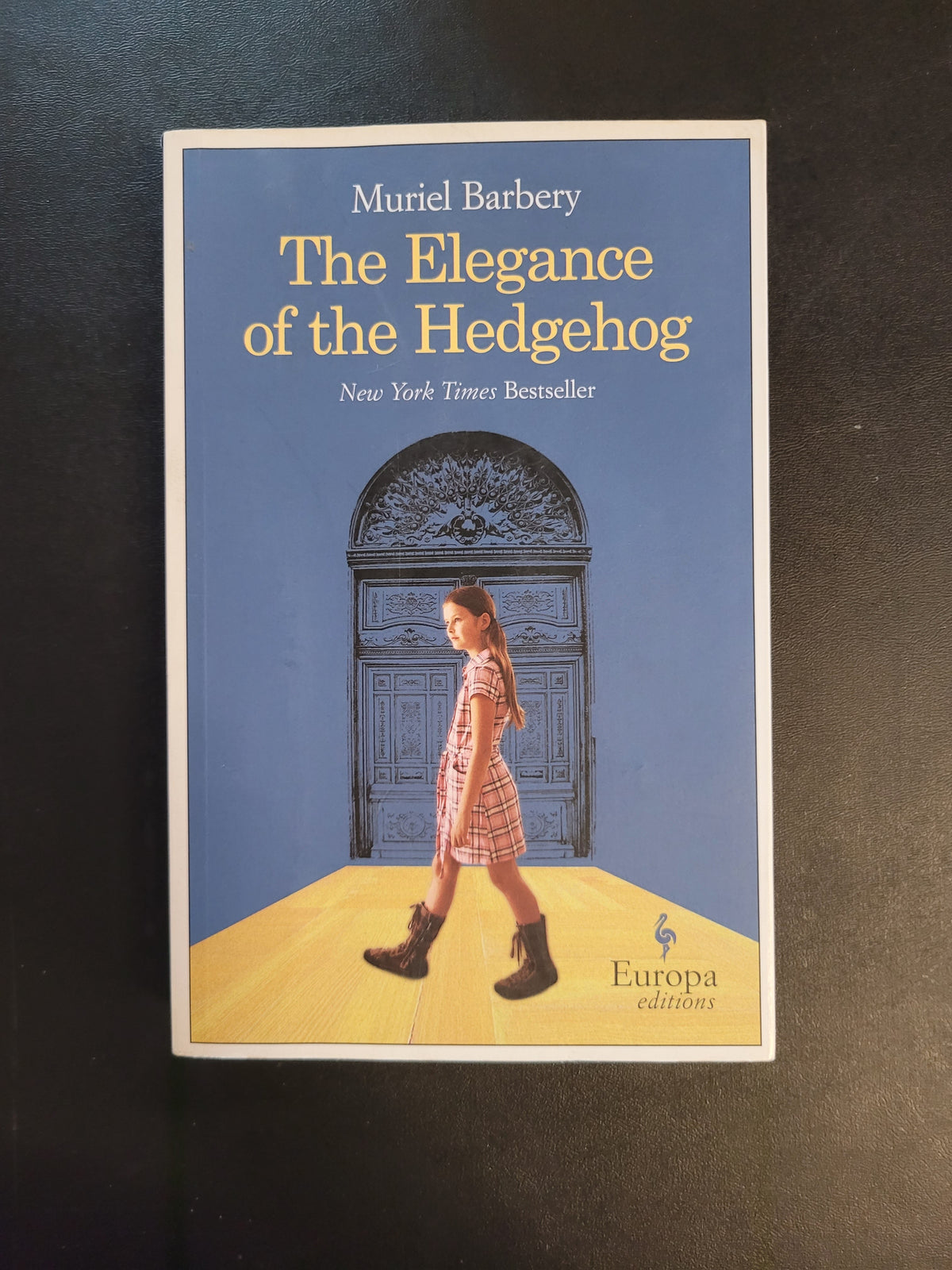 The book cover of 'The Elegance of the Hedgehog' by Muriel Barbery, featuring a blue background, a yellow diagonal section, and an illustration of a girl in a pink plaid dress standing in front of a large door, placed on a dark textured surface.