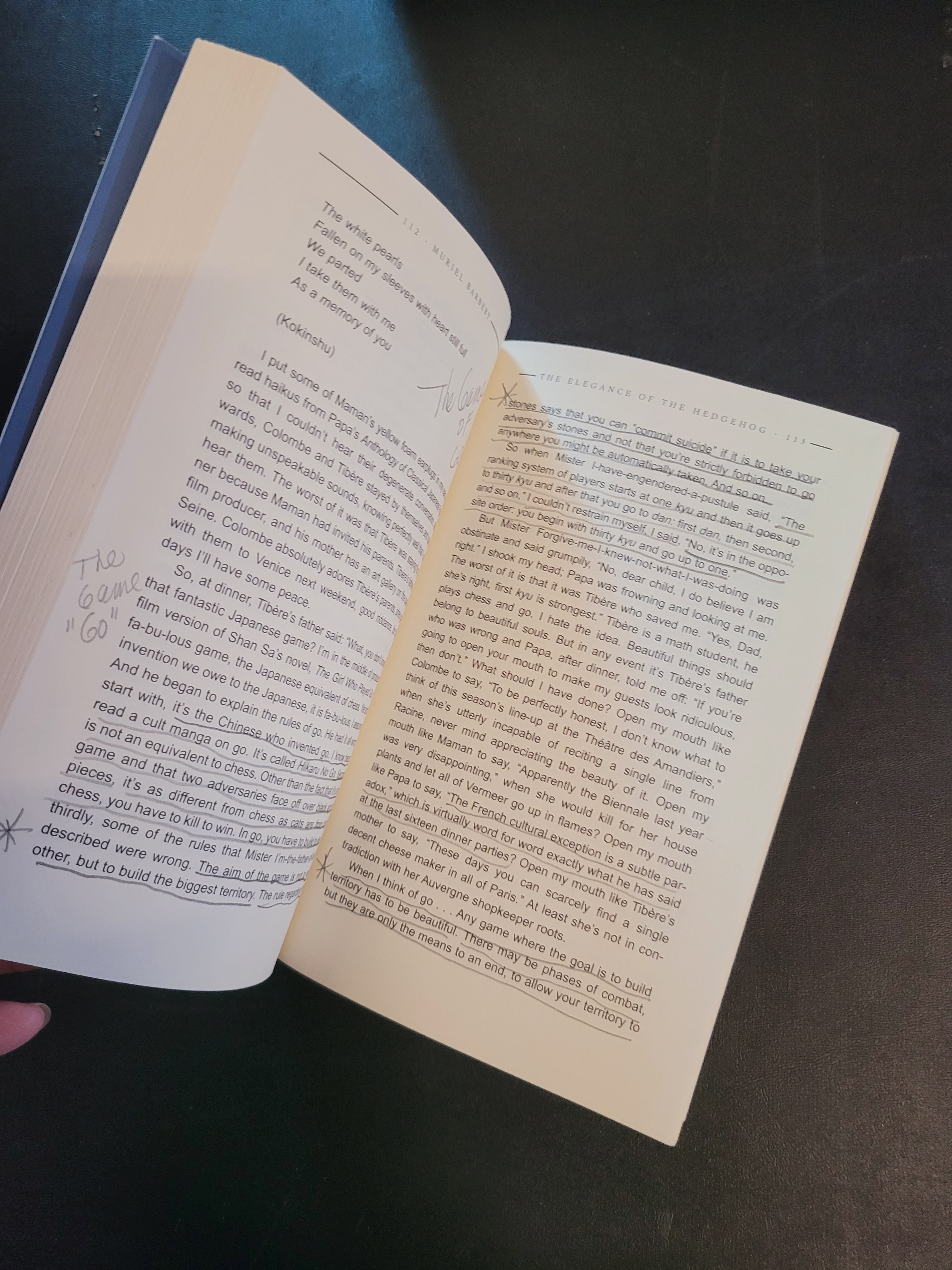 An open book with cream-colored pages filled with printed text, photographed against a dark background. A small portion of a finger is visible at the bottom right holding the book open.