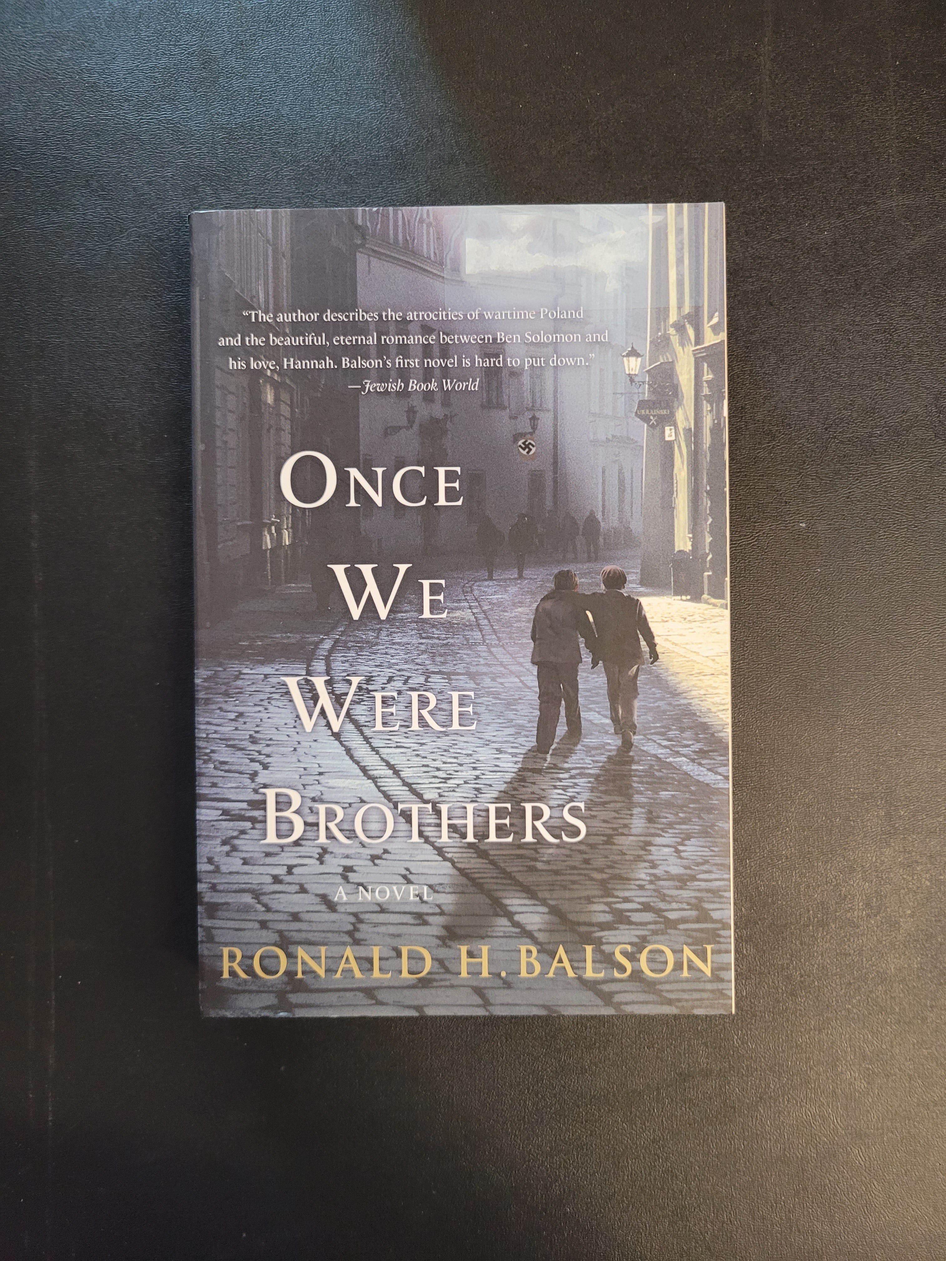 A book titled 'Once We Were Brothers' by Ronald H. Balson, featuring a dark, moody cover illustration of a couple walking on a cobblestone street, photographed on a dark textured surface.