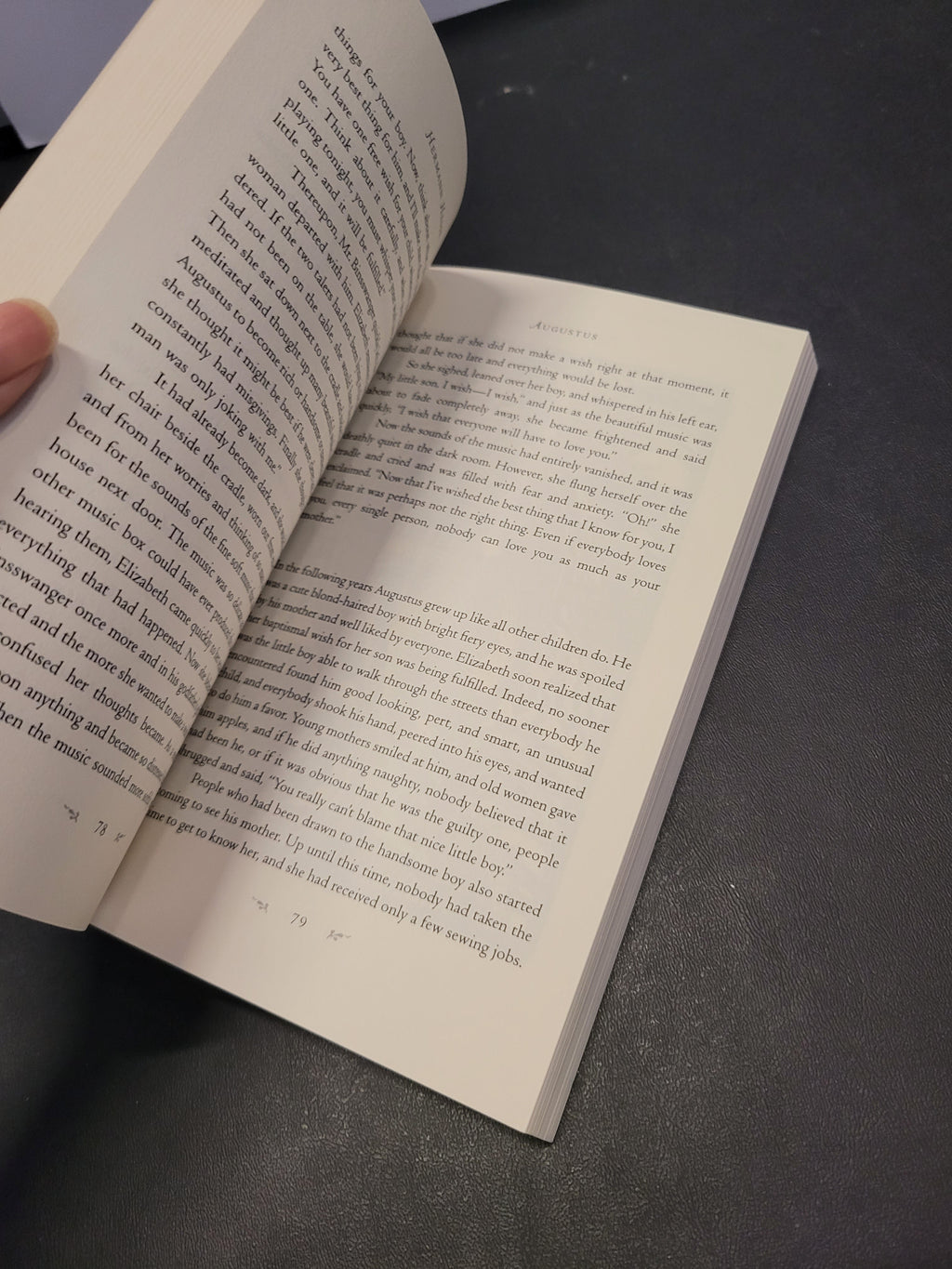 An open book with printed text pages, held slightly by a hand, resting on a dark textured surface.