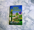 The book 'Hot Shot' by Fern Michaels lies on a white and gray marble surface. The book cover features a desert landscape with a saguaro cactus, palm trees, and a blue sky with yellow title text.