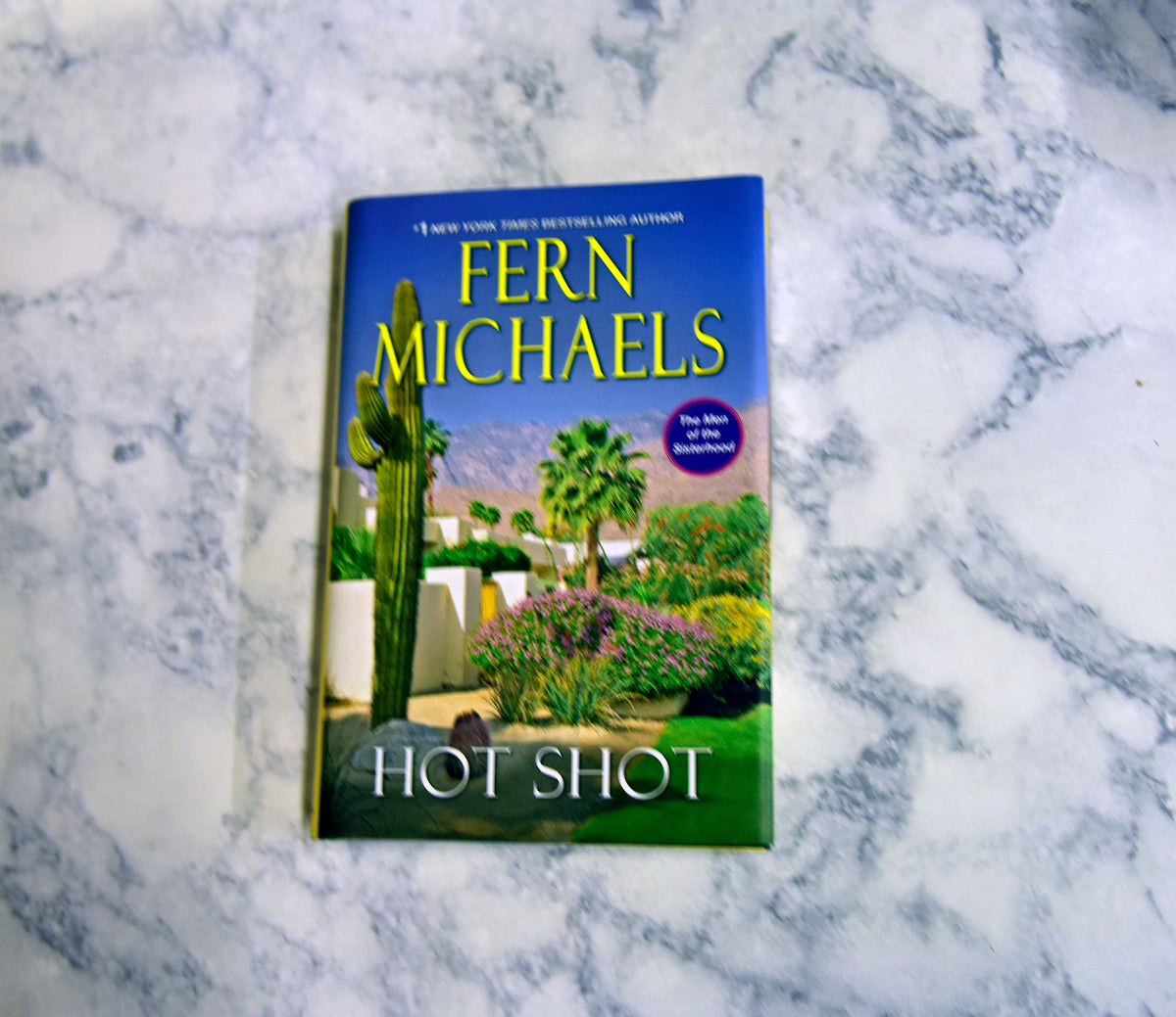 The book 'Hot Shot' by Fern Michaels lies on a white and gray marble surface. The book cover features a desert landscape with a saguaro cactus, palm trees, and a blue sky with yellow title text.