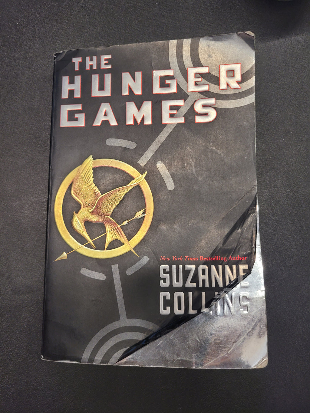 A paperback copy of The Hunger Games book by Suzanne Collins, featuring the iconic golden mockingjay logo on a dark gray cover, photographed on a black background.
