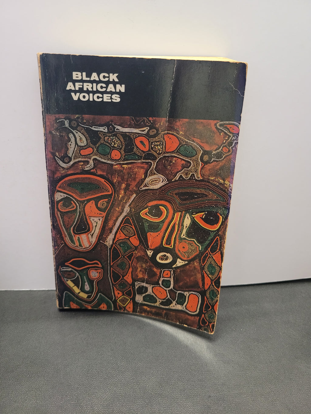 A vintage book titled 'Black African Voices' with a black cover and an abstract painted illustration in orange, red, green, and brown tones, standing on a gray surface against a white wall.