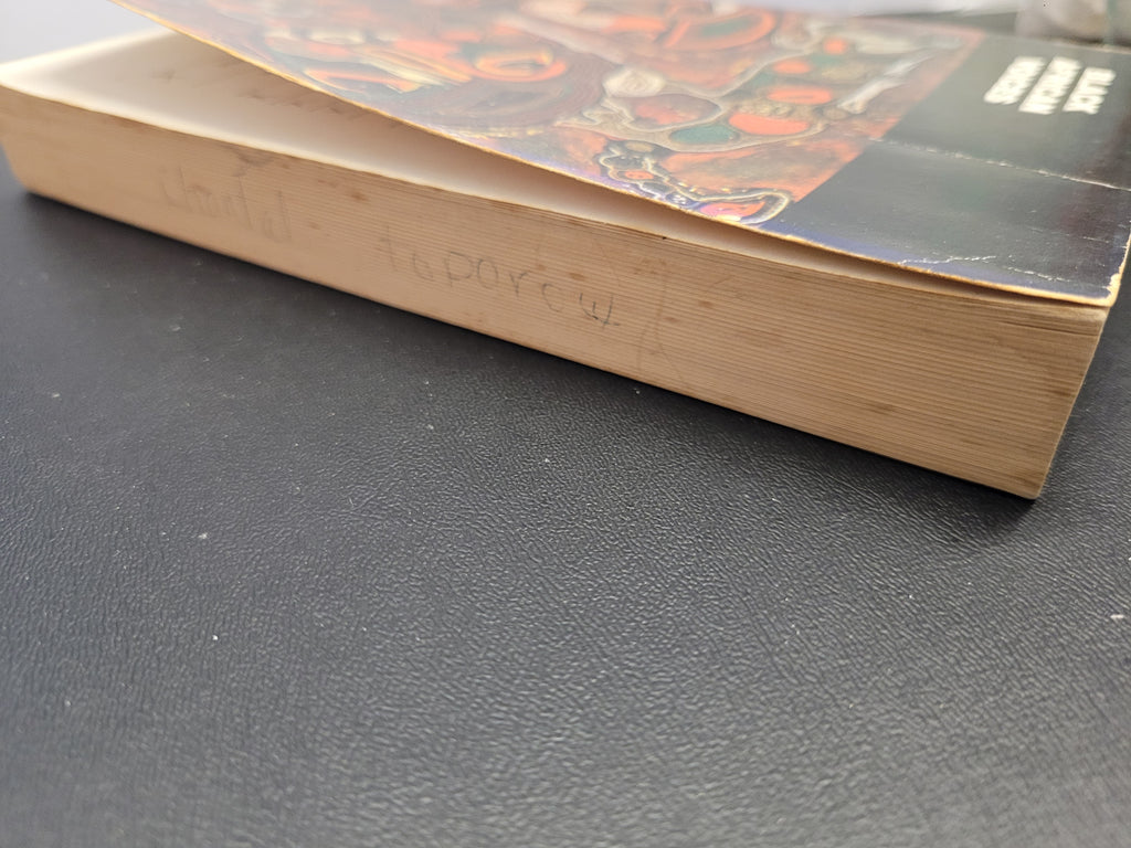 A close-up of a "Black African Voices" book's outer pages and right side of cover resting on a dark textured surface. The pages are light beige with handwritten text, and the cover features a dark background with an abstract multicolor illustration.