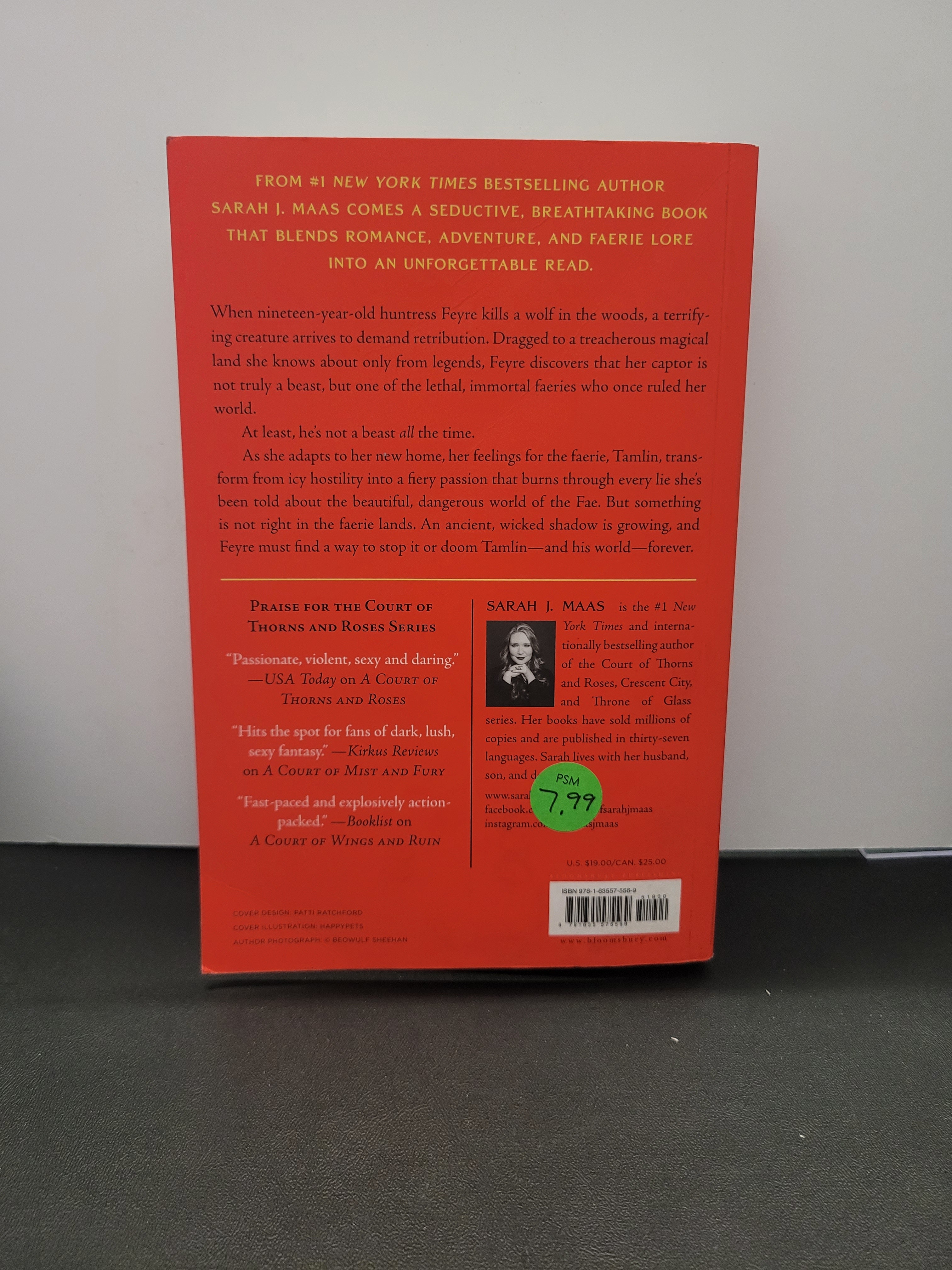 The back cover of a A Court of Thorns and Roses with a bright red-orange background, black text, a barcode, and a small green circular sticker, photographed on a white surface with a dark gray area at the right.