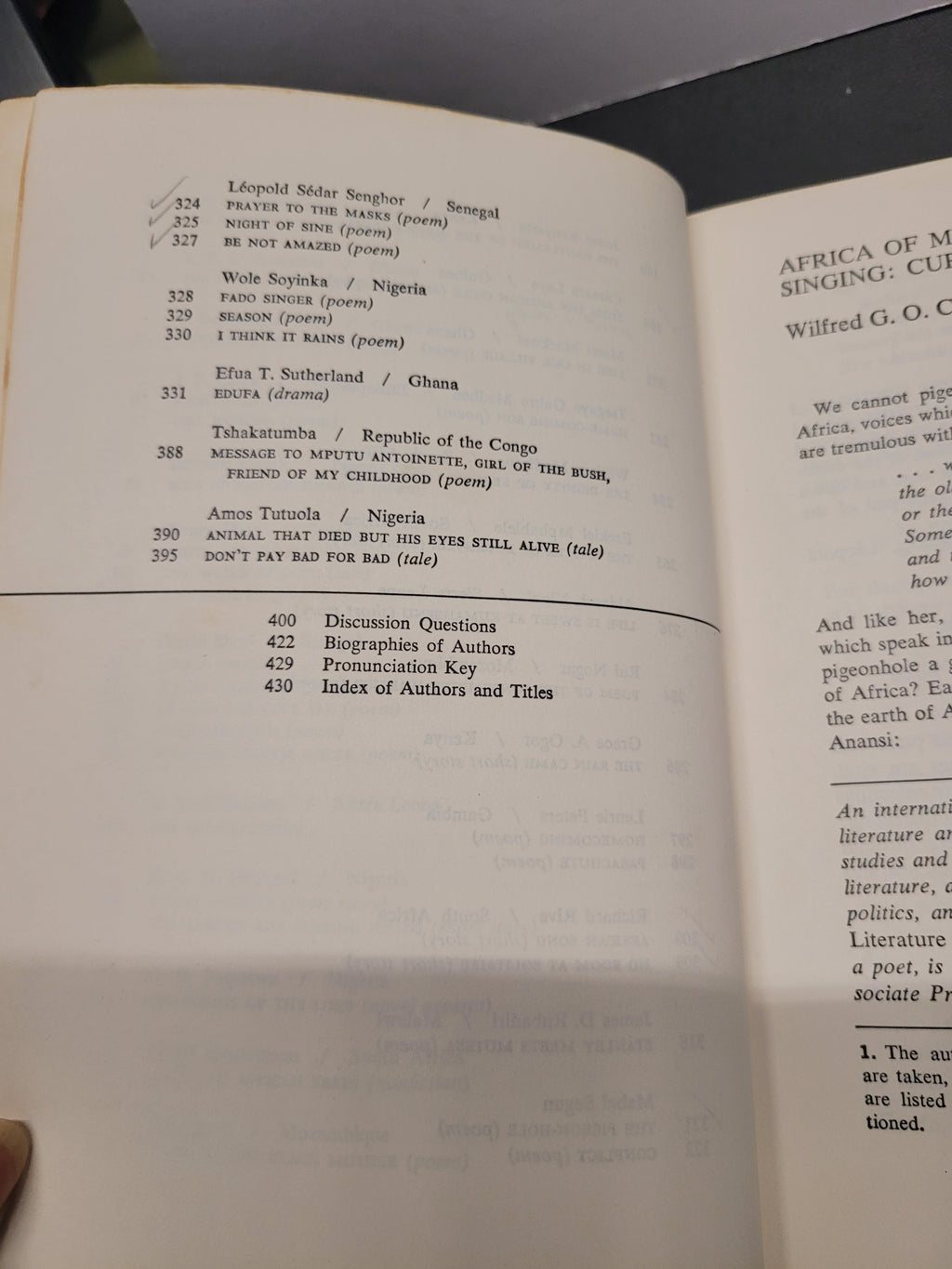 An open book "Black African Voices" showing printed text pages, photographed from above on a dark surface. The left page contains a list of poem titles and authors, and the right page shows a poem excerpt titled 'Africa of My Singsing' by Wilfred G.