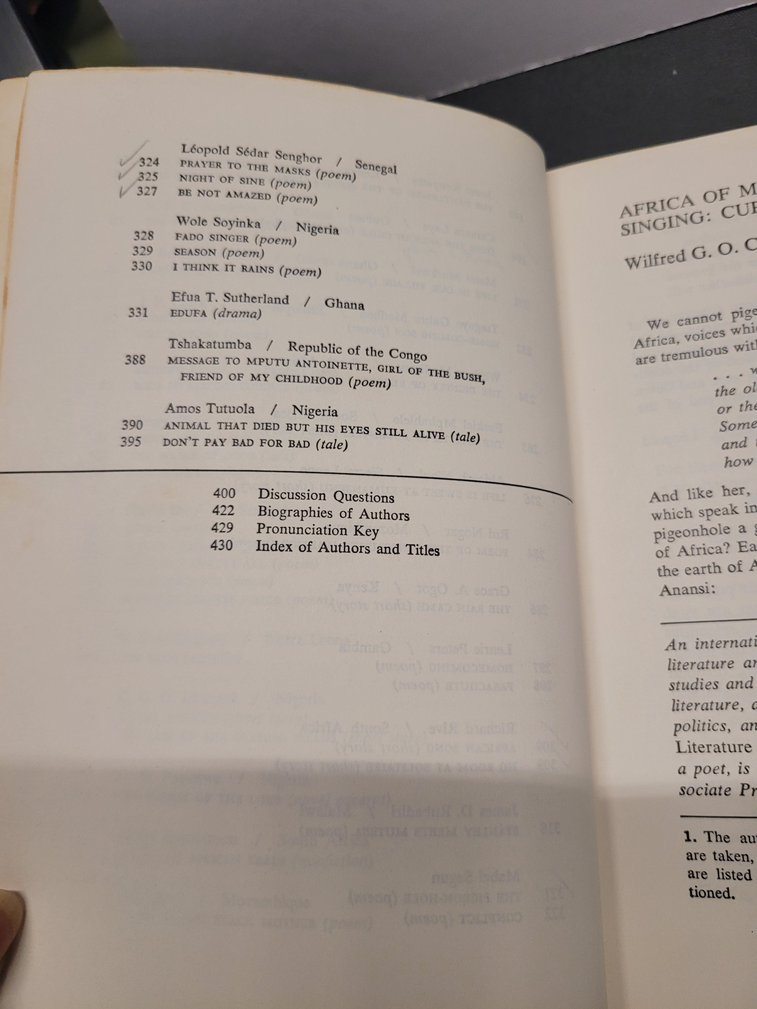 An open book "Black African Voices" showing printed text pages, photographed from above on a dark surface. The left page contains a list of poem titles and authors, and the right page shows a poem excerpt titled 'Africa of My Singsing' by Wilfred G.