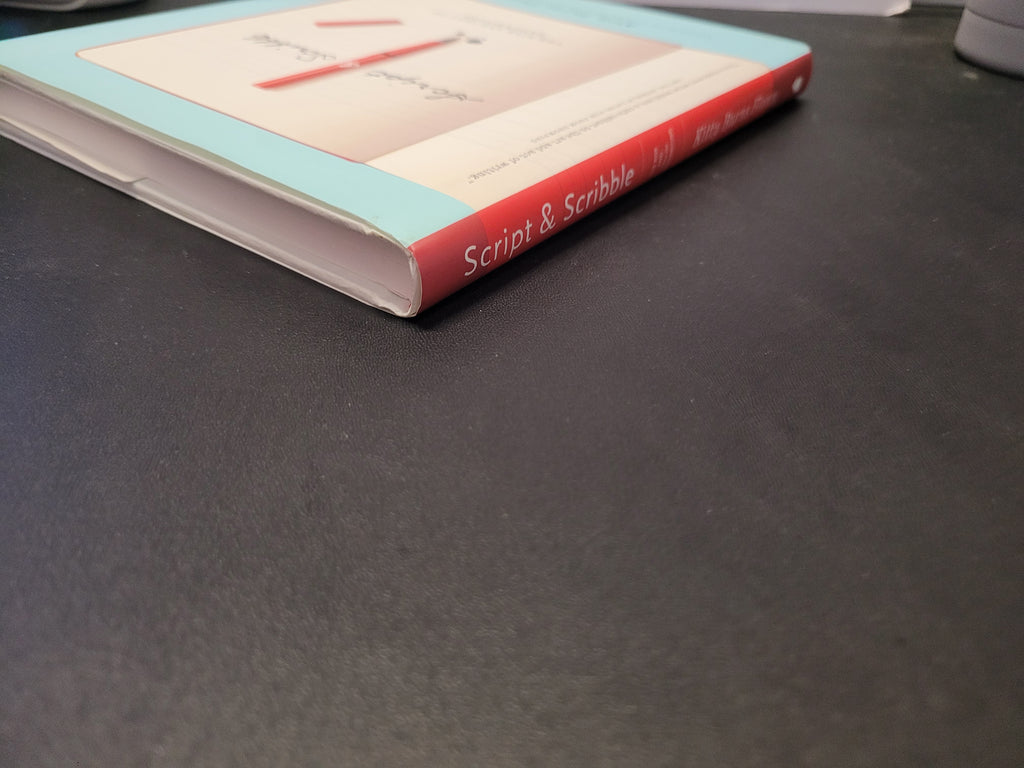 A hardcover book titled 'Script & Scribble Vol. 1' resting on a dark gray textured surface, showing a light blue cover with a red spine.