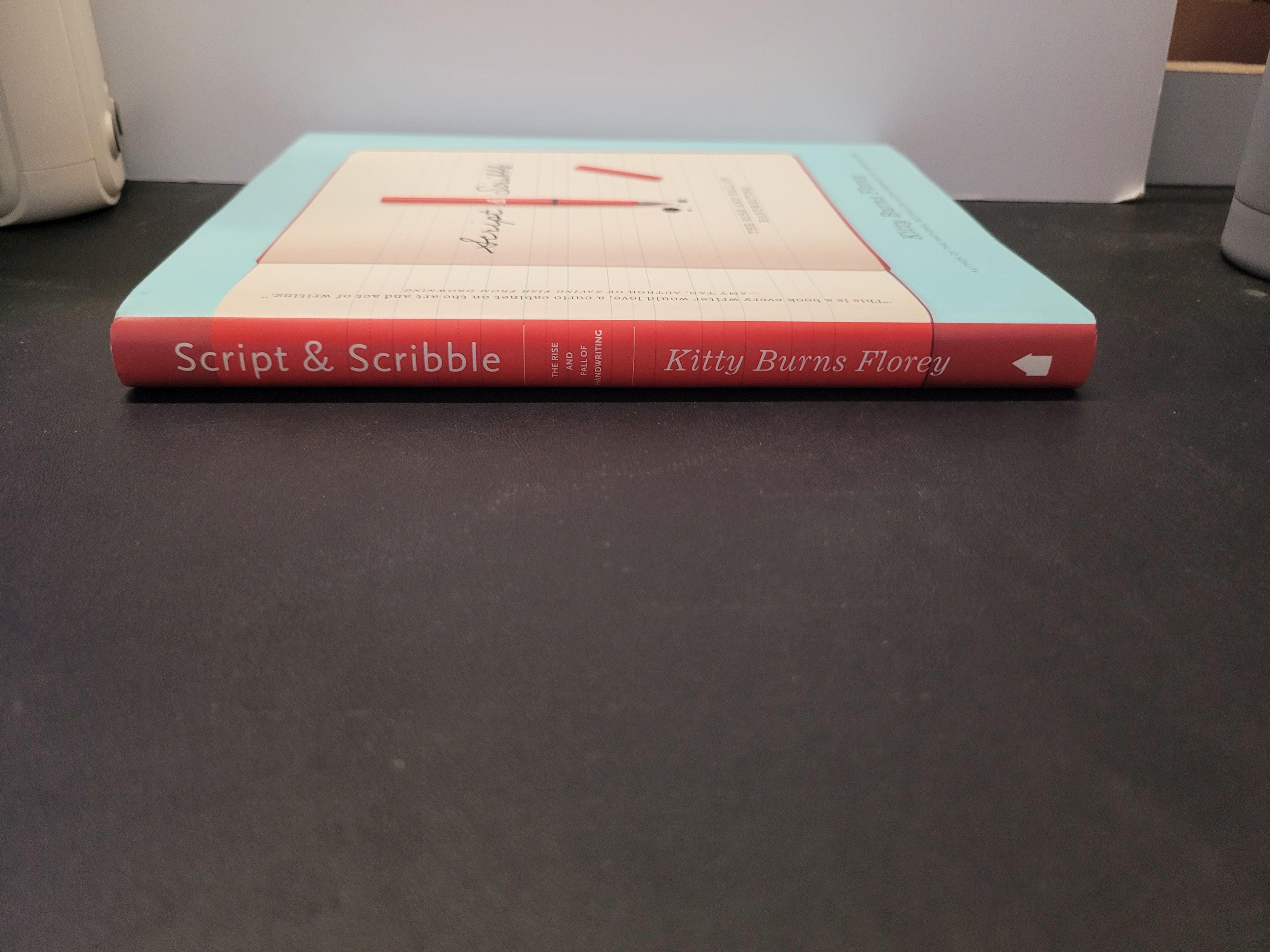 The book 'Script & Scribble' by Kitty Burns Florey, showing its red spine and cream cover, resting on a dark gray surface.